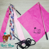 A flat-lay product display of an equestrian tail bag set by Kari's Flags & Tail Bags, featuring a bright pink flag, a patterned pink and multicolor tail strap, a black rope, and a silver telescopic pole on a light wood background.