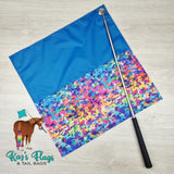 Colorful flag with a blue top and multicolored bottom on a wooden surface, featuring Kaz's Flags & Tail Bags branding.