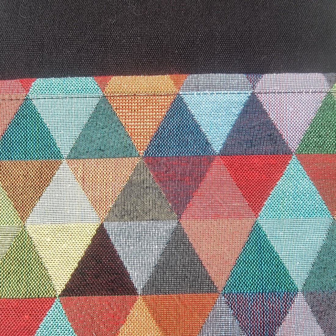 Close-up of a woven textile with a geometric triangle pattern in multiple colors including teal, orange, red, blue, gray, green, and cream, with a black fabric section at the top.