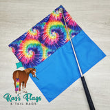 Colorful tie-dye flag with a blue section and a horse illustration, on a light wooden surface with 'Kaz's Flags & Tail Bags' branding.